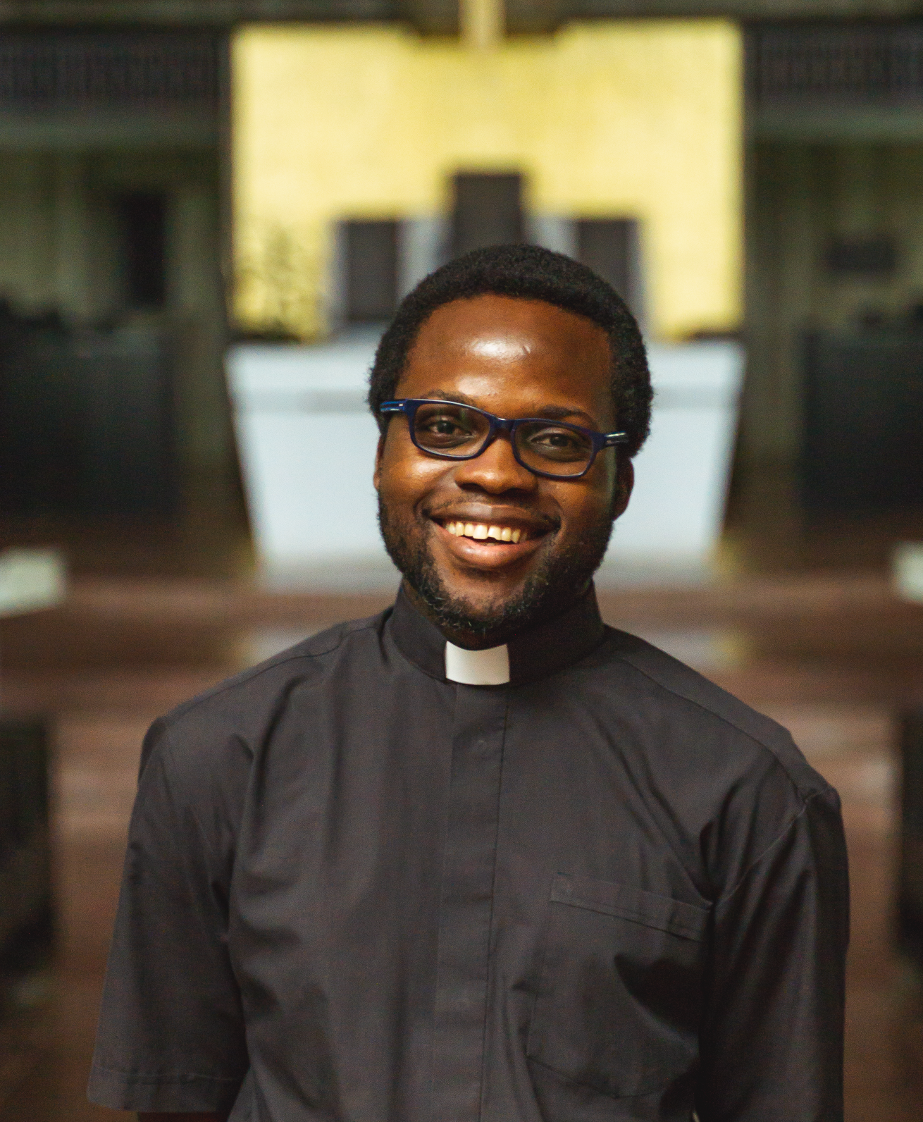 A smiling man wearing glasses and a black clerical collar stands in a dimly lit room with a golden background and dark furnishings.