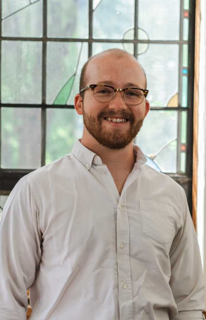 A man with short hair, a beard, and glasses is smiling while wearing a white button-up shirt. He is standing in front of a window with stained glass panels.