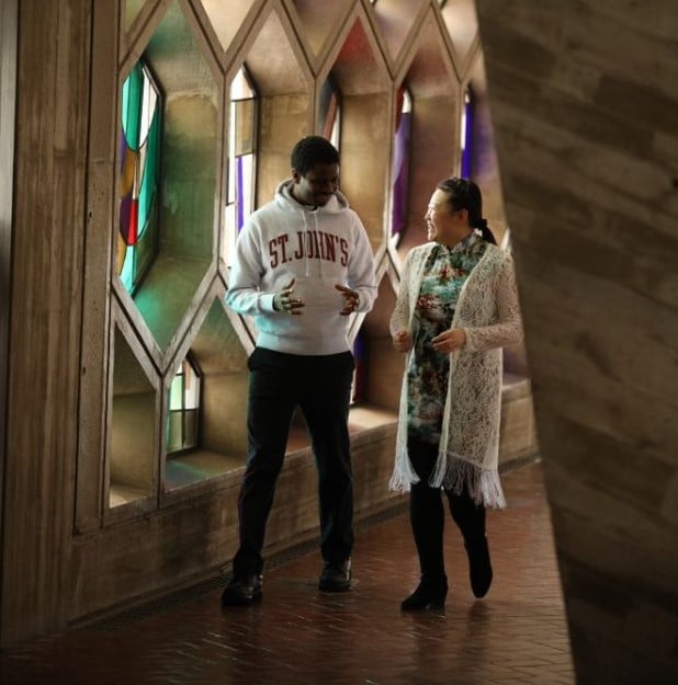 Two people are walking and conversing inside a building with colorful stained glass windows. One wears a "St. John's" hoodie and jeans, and the other wears a floral dress with a white lace cardigan. The hallway features unique architectural elements and a brick floor.