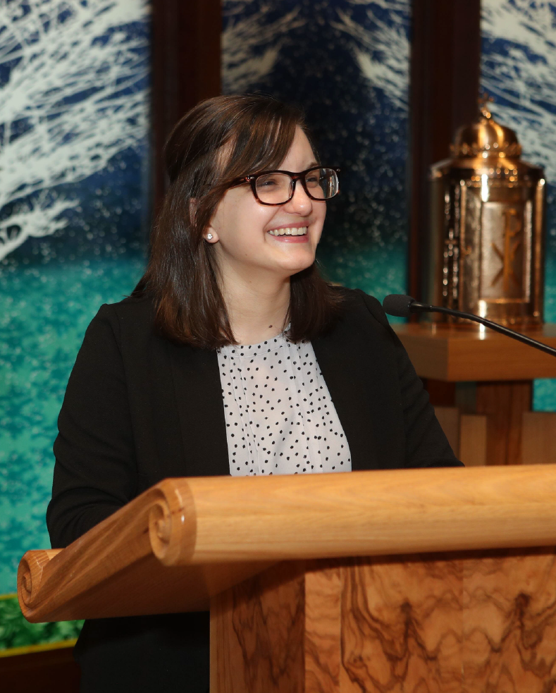 A person in glasses stands at a wooden podium, smiling. They wear a black blazer and a polka-dotted shirt. A decorative background with blue and green tones is visible, along with a large metallic object beside them.