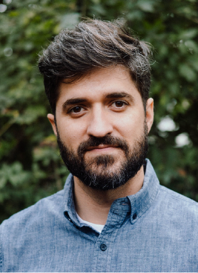 A person with short dark hair and a beard is wearing a blue button-up shirt, standing outdoors with greenery in the background. The individual has a neutral expression.