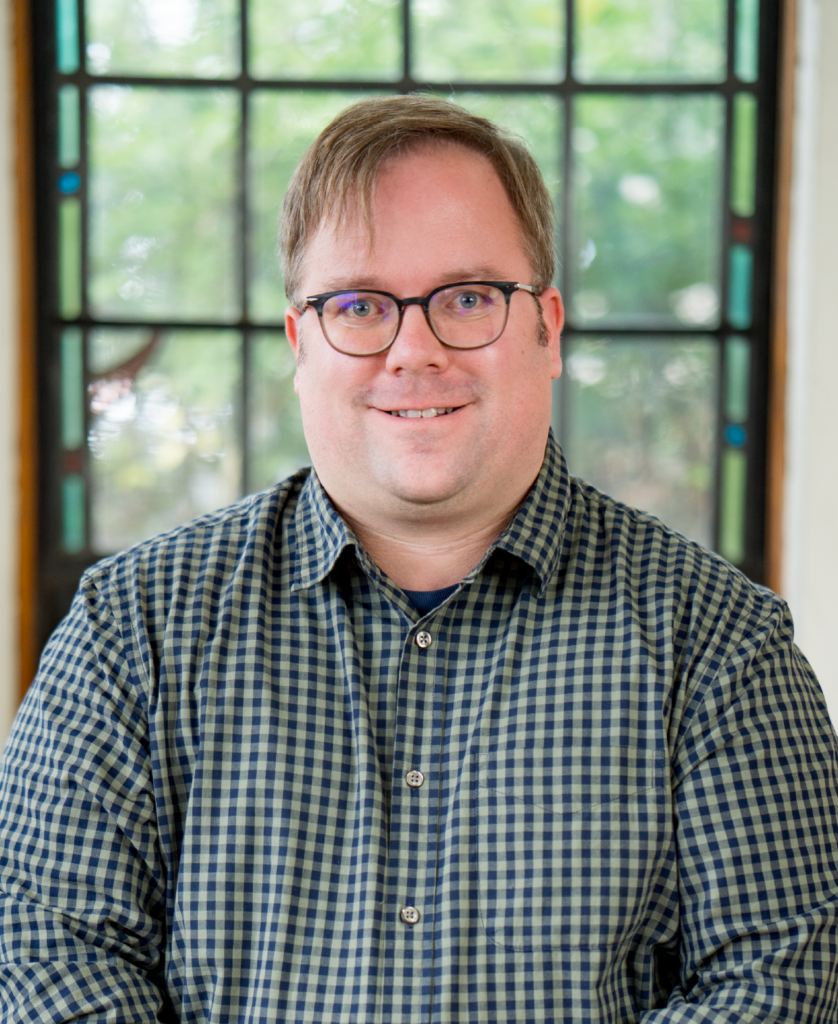 A person with short hair and glasses is wearing a checkered shirt, standing in front of a window with a blurred background of greenery.