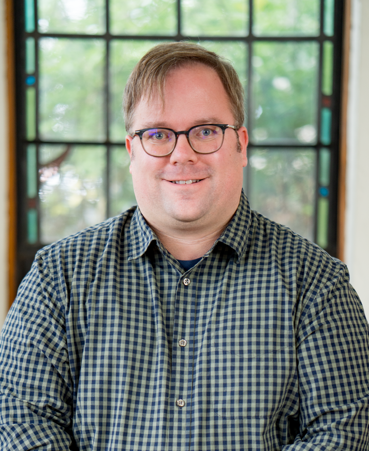 A person with short hair and glasses is wearing a checkered shirt, standing in front of a window with a blurred background of greenery.