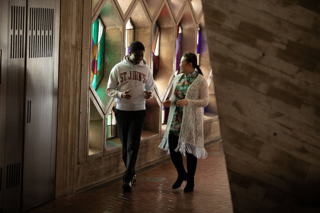 Two people walking and conversing inside a building with colorful stained glass windows on the left. One wears a St. John's hoodie, and the other wears a floral dress with a light cardigan. The floor is made of dark brick tiles.