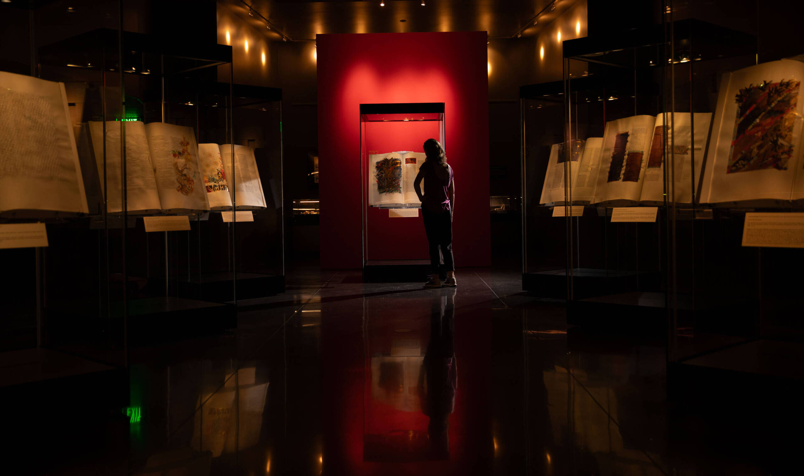 A person stands in front of a brightly lit display of illuminated manuscripts in a dimly lit exhibit hall. The surrounding showcases contain open books with ornate illustrations, creating a dramatic contrast with the red backdrop.