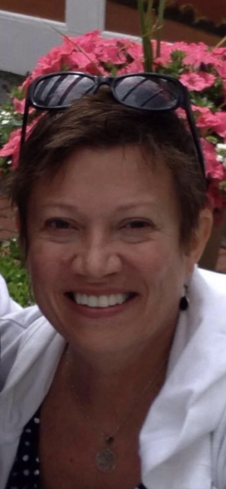 A person with short brown hair smiles at the camera. They are wearing a white jacket and a necklace, with sunglasses resting on their head. Vibrant pink flowers are in the background.