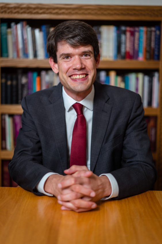 A man in a dark suit and red tie smiles warmly, sitting at a wooden table with his hands clasped. Behind him, bookshelves filled with various books create a scholarly background.