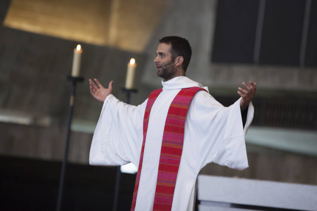 A person in religious attire with a white robe and red stole gestures with both arms outstretched. They stand indoors near two lit candles on tall stands. The background is softly lit and features a modern architectural design.