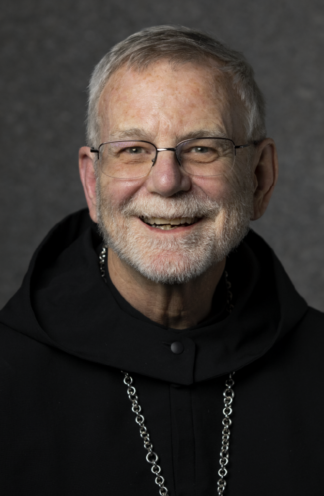 An elderly man with glasses and a gray beard smiles at the camera. He is wearing a black hooded garment and a chain necklace. The background is gray and softly focused.