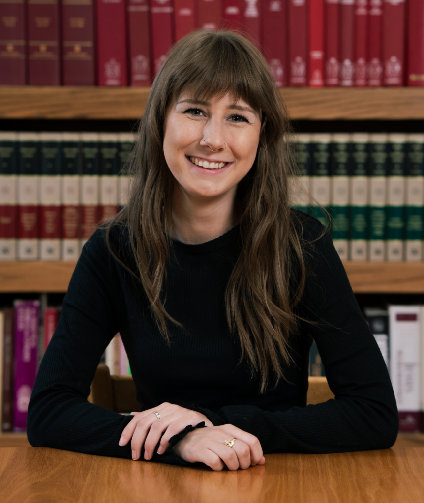 A person with long brown hair and a black top smiles while sitting at a wooden table. Shelves filled with colorful books are in the background.