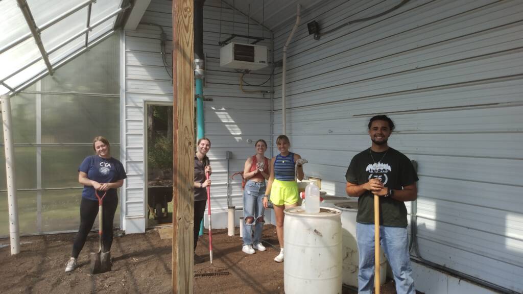 A group of five people stand inside a greenhouse, each holding a gardening tool. They are working on what appears to be a gardening or landscaping project, as the ground is bare soil. The group is smiling and appears to be taking a break from their work.