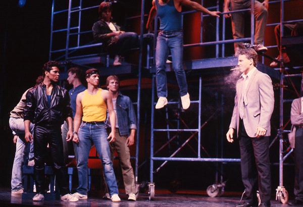 A group of men in a theater production confront one another. Two men in the foreground face each other, one in a leather jacket and the other in a suit. A man jumps in the background, while others watch from scaffolding. Smoking creates dramatic tension.