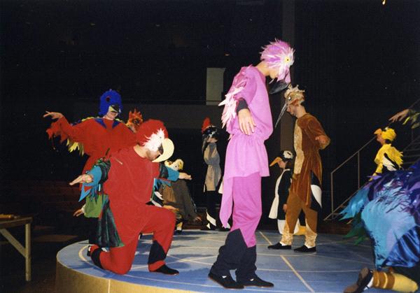 A group of people in vibrant bird costumes are performing on a circular stage. One person in red kneels, while others in colorful outfits pose dramatically. The background is dark, with minimal lighting highlighting the performers.