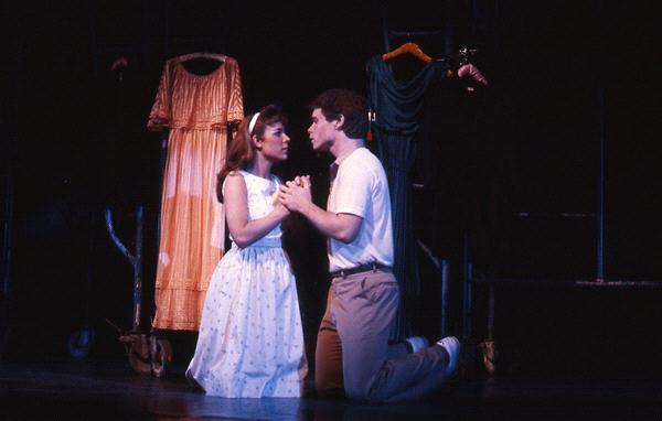 Two actors kneel and hold hands in an emotional scene on stage. Behind them, costumes hang, including an orange dress. The woman wears a white dress with a headband, and the man is in a white shirt and tan pants. The lighting is dim and dramatic.