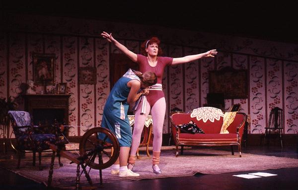 Two women are on stage in a theatrical performance. One woman in a blue dress leans on another woman in a red leotard and leg warmers. They are surrounded by vintage furniture and floral wallpaper in a room setting.