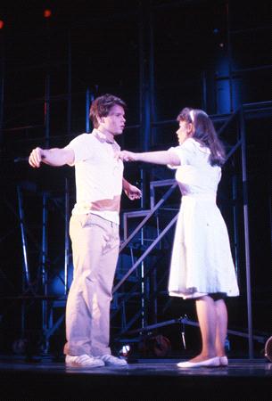 A male and female performer face each other on stage, both in mid-gesture with arms partially raised. They wear light-colored costumes; the man in a shirt and pants, the woman in a dress. The stage backdrop is dark with structural elements.