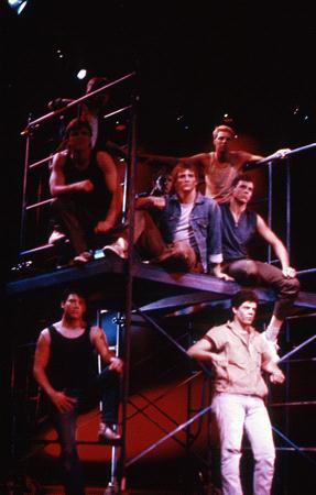 Six men are positioned on a metal scaffolding structure on a stage. They are dressed in rugged, casual clothing, including vests and jeans, with some standing and others sitting. The background is dimly lit, creating a dramatic atmosphere.