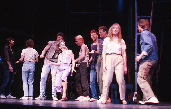 A group of people on stage during a performance. Some are wearing casual 80s-style clothing like jeans and t-shirts. The scene appears lively, with people interacting energetically, and the lighting is dim, emphasizing the characters.