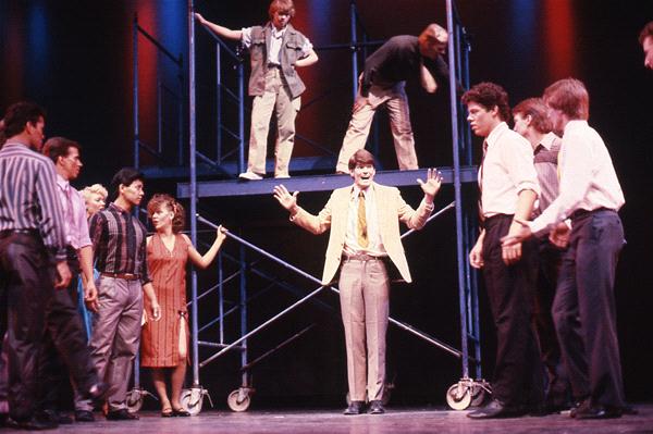 A group of performers on stage, some standing on scaffolding, others on the floor. One person in a light suit gestures energetically in the center. The scene is set with colorful stage lighting.