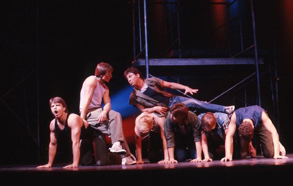 Seven performers are on stage. Six are crouched closely together in two rows, acting as a base, while one is posed on top of them. The scene is dramatically lit with dark structural elements in the background.