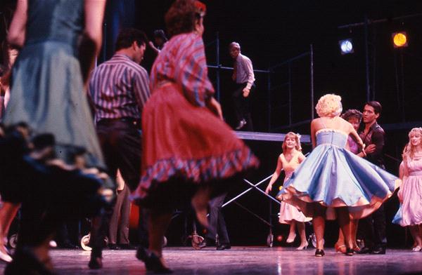 Dancers in colorful 1950s-style outfits perform energetically on stage. Some wear bright dresses with petticoats, while others wear shirts and pants. A man on a platform in the background appears to be observing the scene. Vibrant lights illuminate the stage.