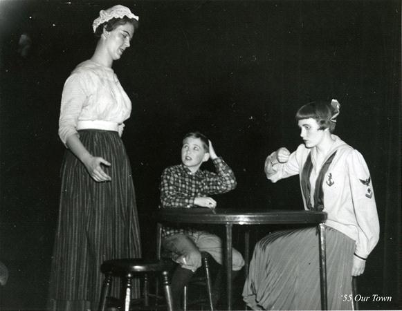 Black and white photo of three people on stage. An adult stands and gestures, while a child and another adult sit at a round table. The seated adult appears to be miming eating. The scene is labeled "'55 Our Town.