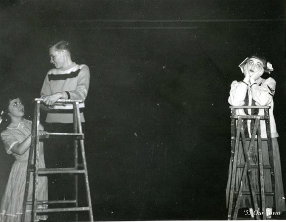 Three actors are on stage performing. Two actors in costumes stand on ladders facing each other, while another actor stands below, looking up and supporting one ladder. The scene is dramatic and the background is dark.