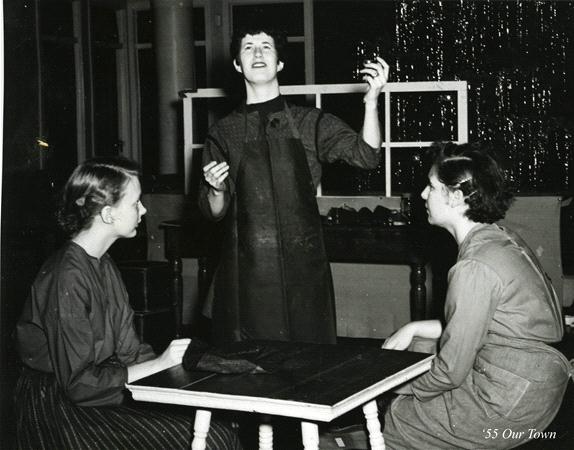 Three people are performing a scene on stage. One stands gesturing expressively, while the other two sit at a table, listening intently. They wear simple, early 20th-century style clothes. The setting is theatrical and dramatic.