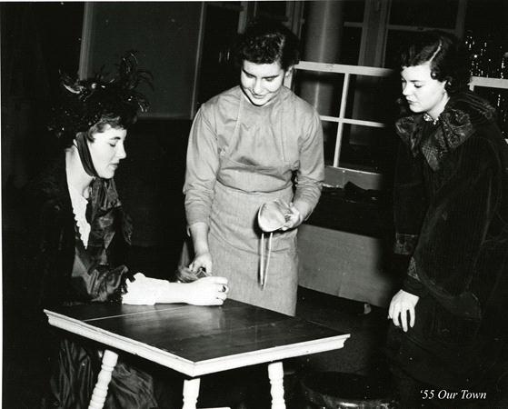 Three people in vintage attire are in a theatrical scene. One is seated at a small table in a feathered hat. The middle person is pouring from a pitcher, wearing an apron. The third looks on, dressed in dark clothing. The text reads "'55 Our Town.