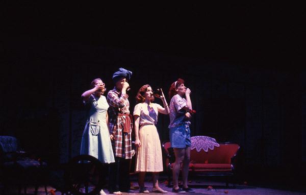 Four people stand on stage in a dimly lit theater, each holding a drink to their mouths. They wear a variety of colorful costumes, including aprons and headscarves. A red sofa is in the background.