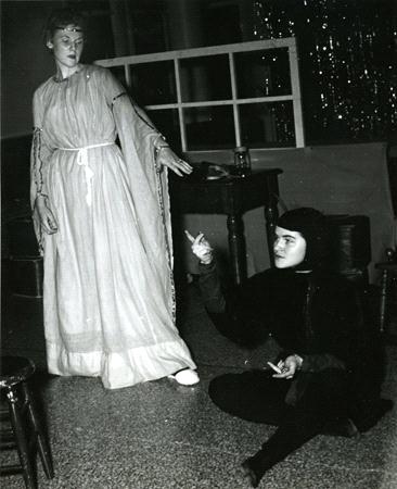 A person in a long dress stands, gesturing towards another person seated on the floor, dressed in dark clothing with a hood. They are in a room with a window and decorations in the background. The scene appears dramatic or theatrical.