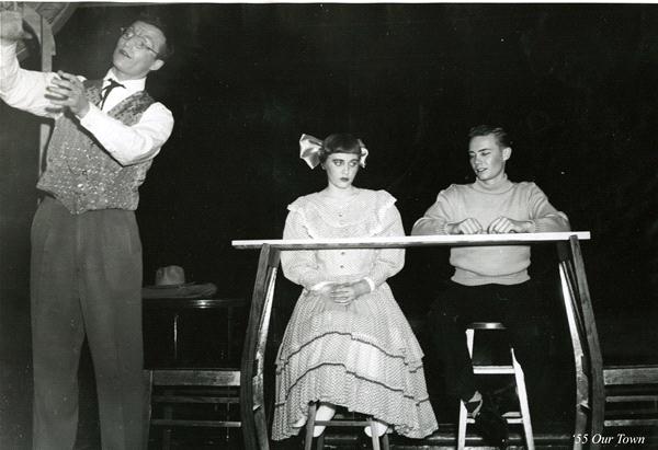 A black and white photo of three individuals performing a scene in a play. A man on the left gestures animatedly, while a woman in a vintage dress and bow sits in the center. A man in a sweater sits on the right, smiling. The set is minimalistic.