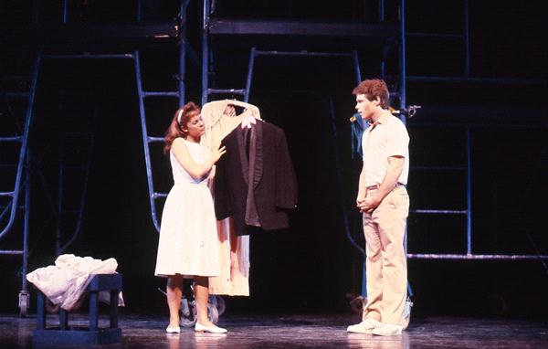 A woman in a white dress holds a dark suit on a hanger, speaking to a man in a light shirt and pants. They are on a stage with a dark background, featuring metal scaffolding and a small table covered with fabric.