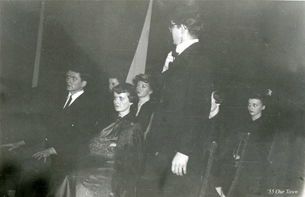 A black-and-white photo of a theater scene from "Our Town" shows a group of actors seated in a semi-circle, listening to a man standing and speaking in front of them. The setting is dimly lit, creating an intimate atmosphere.