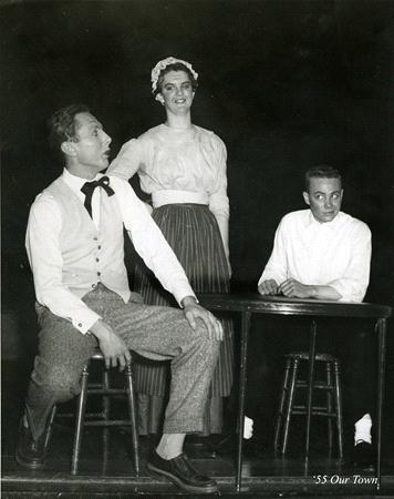 Three individuals in period clothing perform a scene from a play. One sits on a stool, another stands between the seated individuals, and the third sits at a table. The scene has a vintage, theatrical atmosphere.