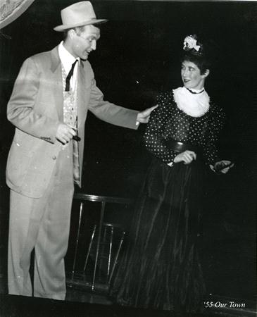 A man in a suit and hat gestures while talking to a woman in a long, dark dress and hat with a white frill, in a theater production. The image has a vintage look, labeled "'55 Our Town" in the corner.