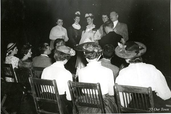 A black-and-white photo of a theater performance. Actors in period costumes stand in a semi-circle, performing. An audience in similar attire is seated on wooden chairs, viewing the scene. The image is labeled "'55 Our Town.