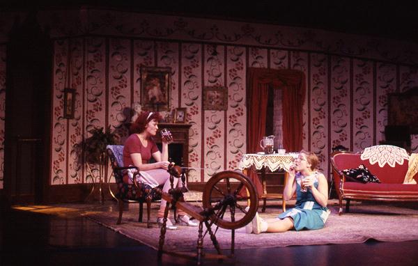 Two actors perform on a stage set as a vintage living room. One sits on a chair, the other on the floor near a spinning wheel, both with surprised expressions. The room has floral wallpaper and period furniture, including a sofa and a lace-covered table.