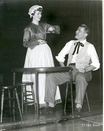 Black and white photo of two people in period costumes on stage. The woman stands, wearing a bonnet and apron, gesturing with her hand. The man sits at a table in a vest and bow tie, looking at her. Stools are nearby. Text reads "'55 Our Town.