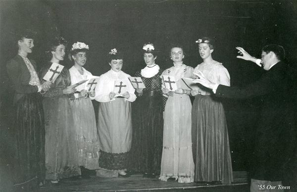 A vintage photograph shows a group of seven women, dressed in long skirts and blouses, holding papers and standing in a line. A man conducts them with his hands raised, also holding a paper. They appear to be singing or performing on stage.