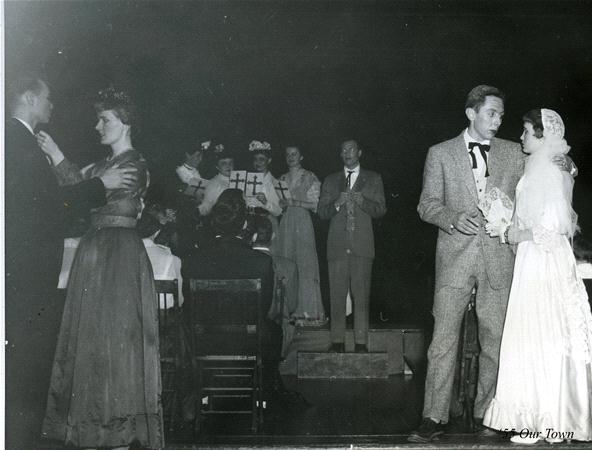 A black and white scene from a theater production with actors dressed in early 20th-century attire. A couple stands on the right, holding hands. In the background, other actors are on stage, with some clapping. The setting suggests a wedding.