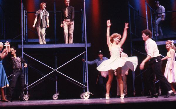 Performers dance energetically on stage with metal scaffolding as a backdrop. One female dancer spins in a white dress, while others are positioned on different levels. The stage is lit with red and blue lights, creating a lively atmosphere.