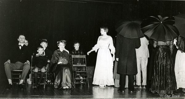 A black and white photo of a theater stage with several actors. A woman in a white dress stands at the center, while others are seated or holding umbrellas. The scene resembles a play set in an earlier time period, labeled "'55 Our Town.