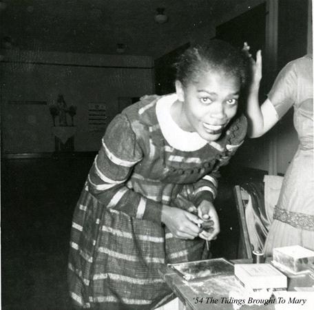 A woman in a striped dress leans over a table, smiling at the camera. She holds a small object in her hands. Another person stands beside her, partially visible. Text in the corner reads, "'54 The Tidings Brought To Mary.