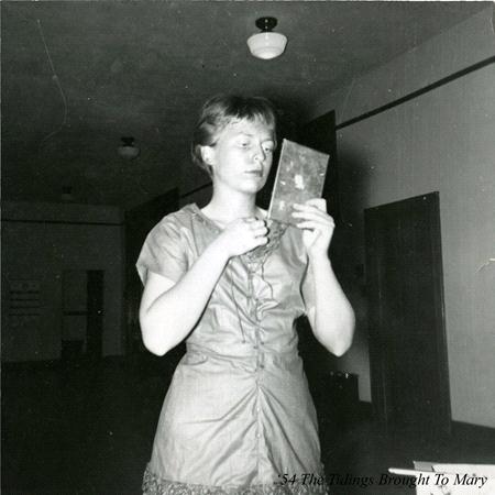 A black and white photo shows a young person standing in a dimly lit room, holding and looking at a small framed object or picture. They are wearing a short-sleeved shirt. The room has plain walls, a door, and a ceiling light fixture.