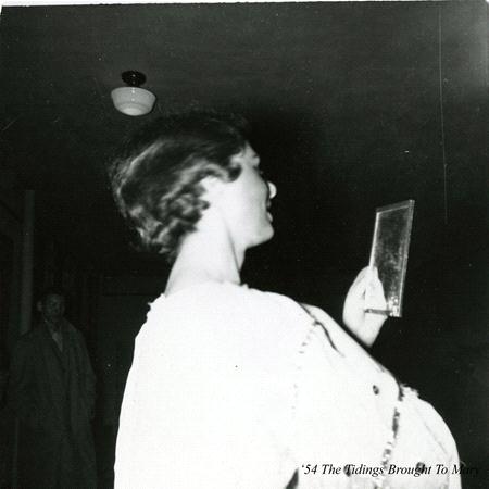 A woman in a white dress poses with a small rectangular plaque or mirror in her hand, smiling in profile. Another person stands in the dim background. The phrase "'54 The Tidings Brought To Man" is visible in the corner.