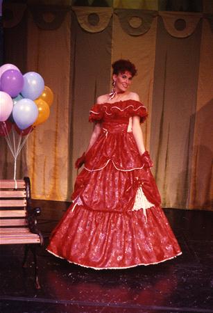 A person stands in a long, red, off-the-shoulder dress with layered ruffles. They are smiling and posing next to a bunch of colorful balloons and a wooden bench. The background features draped curtains.