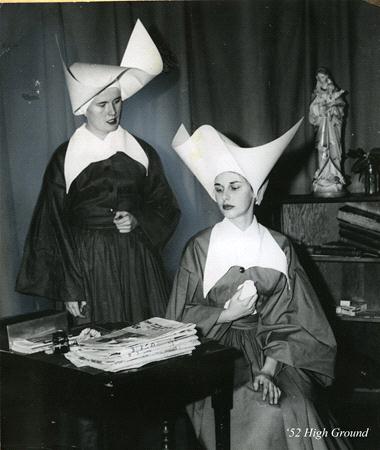 Two women in traditional nun attire with large, pointed white hats are pictured in a room. One is seated, holding a book, while the other stands beside a desk covered with papers. A statue is visible on a shelf in the background.