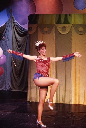 A performer dressed in a circus-style costume, including a tiara and sequined outfit, poses on one leg with arms outstretched, holding a baton. The background features colorful drapes and circular decorations.