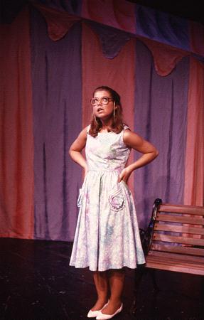 A woman in a light blue floral dress stands with her hands on her hips on a stage. The background features draped fabric in pink and purple hues. A wooden bench is partially visible on the right.